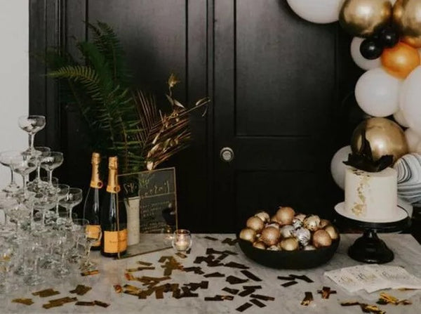Bridal Shower Theme Idea: Brunch and Bubbly. Black doors behind a table full of champagene, empty glasses shaped as a pyramid, gold confetti on the table, and a white cake on a black cake stand. White, gold, black, and orange balloons near the table.