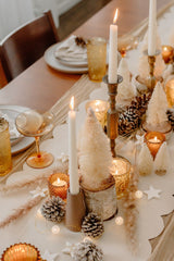 Boho Christmas Table Setting. A cream and soft orange colour scheme. There are white plates and soft orange transparent glasses. A layered table runner with a cream linen, a thick white wavy runner on top, and scattered festive decorations. There are cream brush bottle trees on miniature wooden stumps and white taper candles on vintage candle holders..