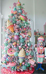 A maximalism Christmas Tree packed with pastel decorations: massive ice creams cones, doughnuts, pieces of cake, glittery bulbs, lolipops, nutcrackers and more. The room is an off-grey colour and has pastel christmas decor off to the side.