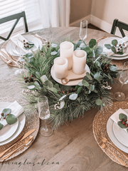 Boho Christmas Table Decor. Rattan table mats with white dishes, gold cutlery, champagne glasses, and berries. The centrepiece is wreath with winter greens, eucalyptus, baby's breath and berries. There are 3 pillar candles sitting on a cream ceramic tray in the middle of the wreath. 