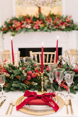 Traditional Christmas Table Setting: white linen table cloth, gold accented plates, gold cutlery, and crystal glasses with a gold rim. The table runner is winter greens like pine, willow pussy branches, red berries, and red taper candles on gold vintage candle holders.
