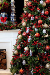 Traditional Christmas Tree decorated with berries, red and gold bulbs, and festive ornaments. The background has a white fireplace full of logs, and a decorated mantel. There are winter greens and red berries as the garland. 