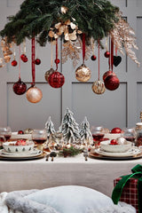 Traditional Christmas Table Setting: an off-white linen table cloth, white plates with Christmas tree details, and the table centrepiece is three miniature frosted Christmas tree sculptures. There is a festive chandelier over the table: pine branches, cream dried flowers, and Christmas bulbs dangling (a mix of red and gold, small and large). 