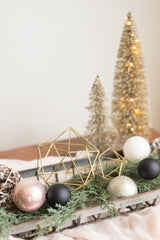 Boho Christmas Decor. A wooden tray with two gold geometric stars, and pink, black, and white Christmas bulbs. There are two frosted brush bottle trees behind it.