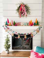 A merry and bright Christmas mantel: a white fireplace with a mini potted tree beside it. Two rainbow pom pom garlands and a wooden christmas tree garland hang across the mantel. Colourful Christmas tree sculptures and a "merry" sign are on top. There is a minimalistic wreath with bells on the wall.