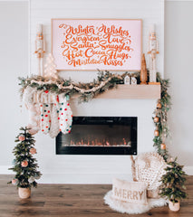 Boho Christmas Mantel. Minimalistic living room with hardwood floors and a glass fireplace with miniature Christmas trees on both sides. The trees are decorated with pale pink ornaments. There is a small faux fur white rug with a "merry" pillow on it.  The white mantel has pale pink and white stockings with coloured pom poms hanging. There is a garland of frosted winter greenery and a garland of beige beads. There is a white, tall nutcracker on each side of the mantel and small festive sculptures. There is a pink sign hanging on the wall  with orange/red letters that say "Winter wishes, evergreen love, Santa snuggles, hot cocoa hugs, mistletoe kisses" 