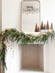 Woodland Christmas mantel decorations: a fresh garland of pine is draped over the white mantel with fairy lights intertwined,  there is a cluster of clay, and there are 6 brown, clay Christmas tree sculptures and a gold mirror on the mantel.