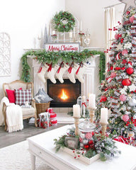 Traditional Christmas decorations in a white living room. There is a centrepiece on the coffee table with large pillar candles and greenery.  A nicely decorated tree with a red/white colour scheme. A white mantel with a fire going: white stockings hanging, a garland of pine, a merry christmas sign on top with a wreath behind it. Other traditional Christmas decor is scatted around the room.