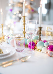 A Merry and Bright Christmas Table Setting: a white table cloth, gold cutlery, white plates with pearl detailing, and gold candle holders with white tapper candles. The table runner is an assortment of colourful Christmas bulb (purple, pink, teal, pastels).