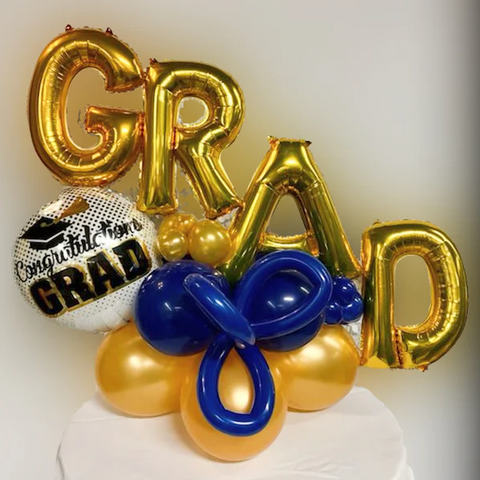 Grad centerpiece made by Party Stuff. There is a gold GRAD foil balloon and a silver foil balloon that says "Congratulations GRAD". There are blue and gold latex balloons at the base.