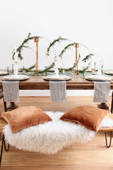 Boho Christmas Table Setting. A minimalistic room with hardwood floors, white walls, and an oak dining bench. The bench has white faux fur and two copper pillows. There are white and grey striped linen napkins, cream coloured plates, and gold cutlery. On each plate is a white tree sculpture with a gold star. The centrepiece is four gold hoop wreathes - all different sizes  - with winter greens and two have a brown linen ribbon.