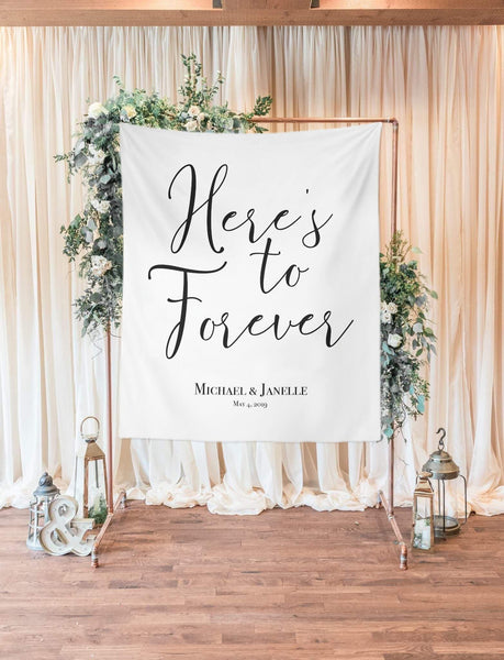 Empty room with a bridal shower backdrop. Gold arch decorated with white/blue flowers and greenery. Candle lanterns on the side. White backdrop on gold arch that says "Heres to Forever" with the name of the couple 