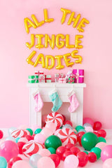 A bright pink room with a white mantel. Over the mantel there are gold balloons saying "all the jingle ladies". On the mantel are gifts with pink wrapping paper. There are three pastel stockings hanging. There are green, pink, and red balloons all over the floor with peppermint candy decor.