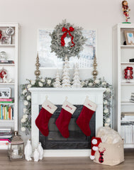 A traditional Christmas matel: white fireplace with three red stockings hanging, a frosted garland hanging off the edges, white Christmas tree sculptures on top, and a Christmas wreath hanging above.