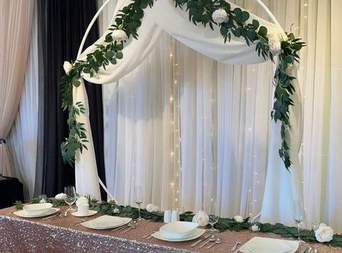 The head table at a wedding with a white backdrop, and an archway with fake vines in front of it. The head table has a pink glittery table cloth with white dishes placed on top. 