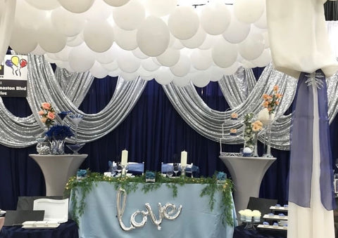 Taken at the Wonderful Wedding Show. A wedding head table set up with silver, glittery drapes and navy blue backdrop. There is a silver "love" balloon attached to the front of the table and a ceiling of balloons above. 
