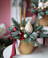 Traditional Christmas Centrepiece: gold pot with frosted pines, two gold taper candles, a deep red ribbon, small bubls, and a painted pine cone. 