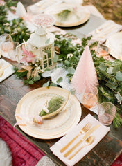 Boho Christmas Table Setting. Light gold and crystal plates, gold cutlery, and off-pink glasses. There is a garland table runner with pink brush bottle trees.