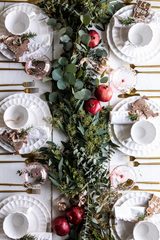 Traditional Christmas Table Setting: white linen table cloth and napkins, ceramic white plates and cups,  Christmas cookies, and a winter greens table runner (eucalyptus, baby's breath, and apples). 