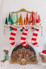 Merry and Bright Christmas Mantel: a white mantel with candy cane stockings, a pom pom rainbow garland,  colourful brush bottle trees, and a gold mirror behind it. Near the fireplace, there is a stuffed reindeer toy.