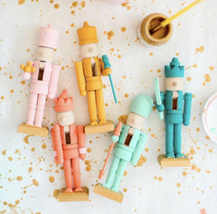 Five pastel painted nutcrackers laying down on a white table with gold paint splatter.