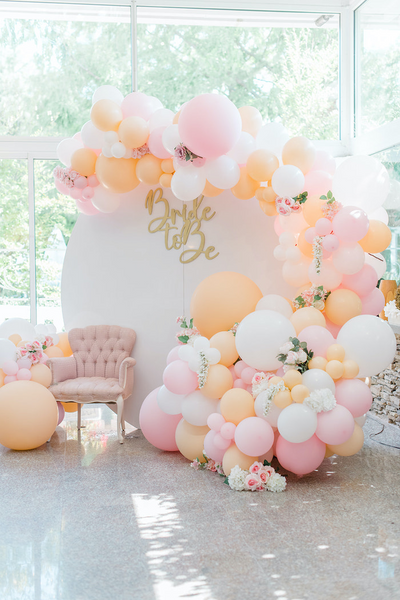 Bridal shower backdrop with pink, white, and orange balloons and garland. Backdrop as a sign "bride to be". Flowers scattered throughout the balloon garland.