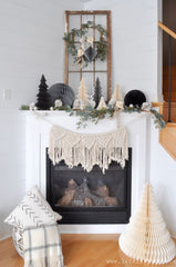 Boho Christmas Mantel. A white fireplace with a basket of pillows and an accordion cream tree near the bottom. There is a macrame garland, and a garland of eucalyptus and other winter greens.  There are white, cream, and black accordion trees on top with small silver bulbs. A repurposed wooden window frame is behind everything with a gold hoop wreath hanging from it. The wreath has winter greens and accordion ornaments.  