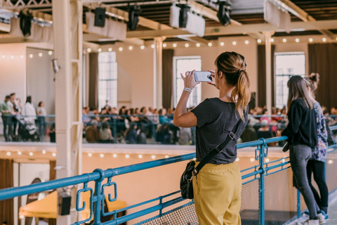 Woman at a venue, in front of a rail, taking a picture. A group of people can be seen in the background