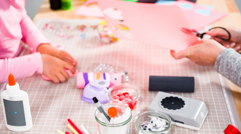 2 young girls hands can be seen doing doing crafts at a table. Pom poms, glue, stamps, scissors, and a toilet paper role made to look like a worm, are on the table.