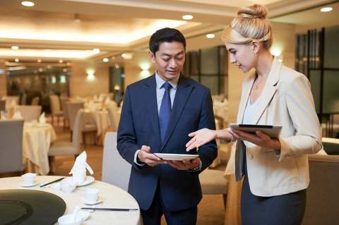 A man and a woman stand in a venue room with nicely decorated tables. The man has a plate in his hand and the woman has an iPad.