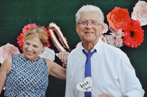 An older woman and man stand in front of a photo booth wall. The wall is green with paper flowers. She is laughing and he is smiling, while holding a small heart sign that says, "Love You".