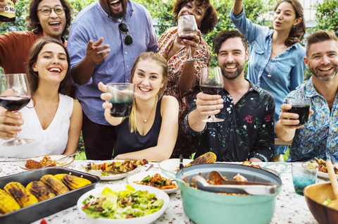 A group of men and woman sit in front of a table full of food. There are 4 people sitting and 4 people standing behind them. Everyone is "cheers-ing" the camera with wine glasses, while smiling.