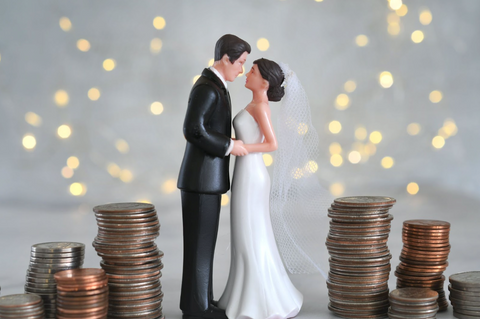 A figure of a bridge and groom kissing, presumably a cake topper. There is money, in the form of coins, piled up on either side of the figures. There are fairy lights in the background.