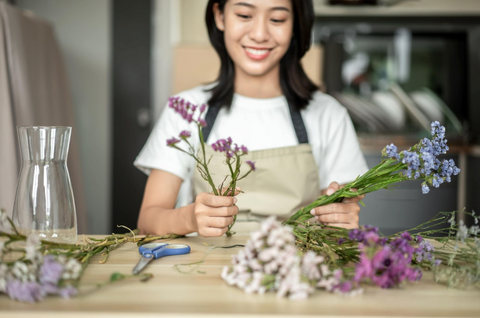 A woman smiles as she puts together a bouquet of wild flowers. There are flowers, scissors, and a vase on the table in front of her.