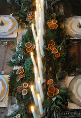 Woodland Christmas Table Setting: a dark wooden table with a garland of pine as the table runner. The garland has dried orange slices and leaves intertwined. There is a row of white taper candles between the garland. There are simple white ceramic plates and glasses with gold rims. 
