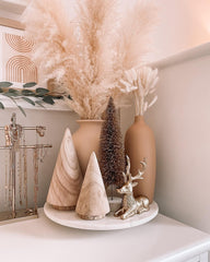 Boho Christmas Mantel Decoration: a marble tray holding two wooden Christmas tree sculptures. The sculptures are smooth and cone shaped. There is a frosted brush bottle  tree and a gold reindeer in a sitting position beside the sculptures. A tanned clay vase is behind the reindeer with cream bunny tails. A large beige clay pot is behind everything with large pampas stems. 