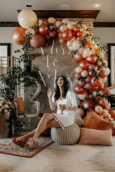 Boho styled room. Autumn coloured balloon garland running up the wall. Miss to Mrs backdrop. Large pillows and chairs. Bride to be in a white dress holding a win glass with a flower crown sitting in front of the backdrop.