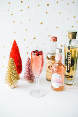 A Merry and Bright Christmas Table Setting: Christmas cocktails. Three brush bottled trees beside a champagne glass with rose and berries. Three alcohol bottles are beside the glass. The background is all white with gold sparkles.