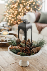 Woodland Christmas Table Setting: a pine cone centrepiece with twigs and pine greenery in an off-white ceramic vase.  It's sitting on an oak table with a black candle lantern behind it.