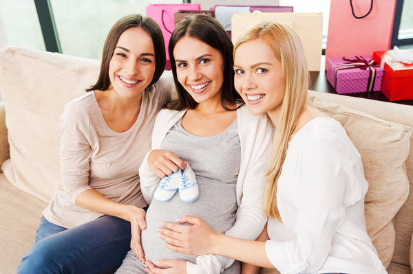 3 women smiling while the women in the middle holds blue baby shoes on her pregnant stomach