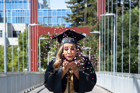 Woman on bridge walkway, in a black graduation gown and cap, blows confetti out of her hands, towards the camera.