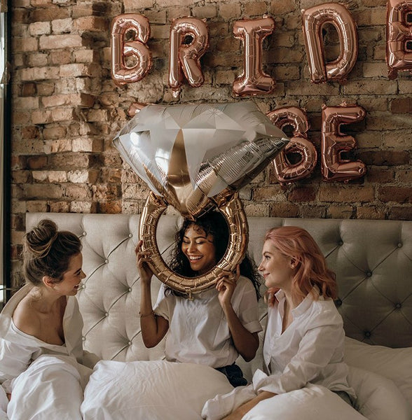 Bridal Shower Theme Ideas: Glam Slumber Party. "Bride to Be" balloon banner on a brick wall. Three women on a bed laughing. The woman in the middle is holding an engagement ring balloon.