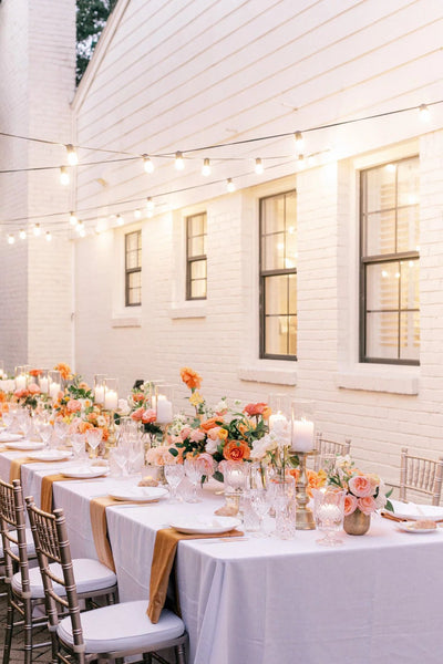 Gold string lights over a bridal shower table: white table cloth, pink and orange flower arrangements, and candles. Outdoors.
