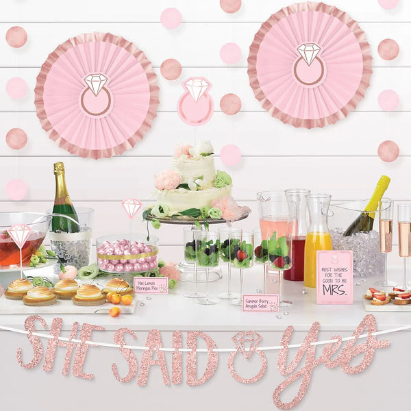 Blush bridal shower theme from Party Stuff: white table full of food and drinks, a pink 'she said yes' banner hanging from it, other blush decorations like wall-hangers and pink cards