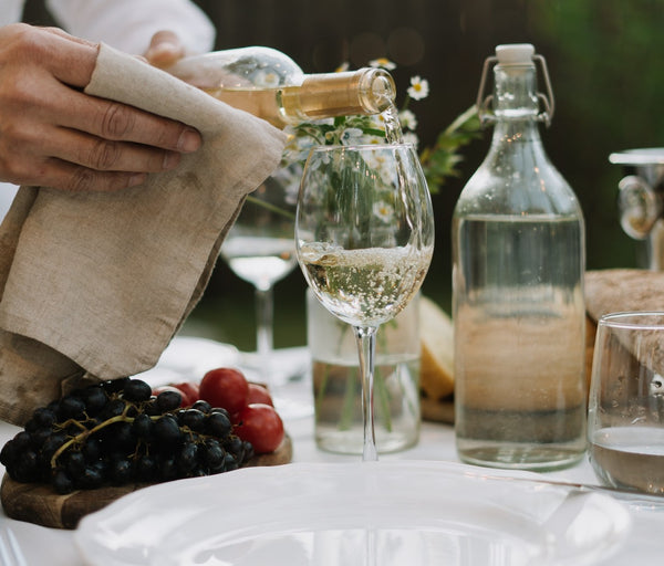 Bridal Shower Theme Ideas: Vino before Vows. A server pouring white wine into a wine glass. Elegant table setting with white linen, a white plate, a fruit board, a glass jug of water,  and bread behind the glasses.