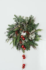 A Christmas wreath hanging on an off-white wall. The wreath is made of pine, eucalyptus, and twigs. There is a string of red ornaments hanging.