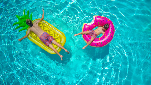 Two young boys on pool floaties in a large pool. One boy is laying on a large pineapple floatie. The second is sitting in a pink donut shaped floatie. The water is very blue. 
