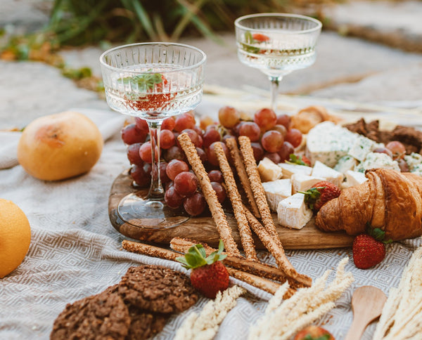 Bridal Shower Theme Ideas: Vino before Vows. Outdoor wine tasting. On a beach with a tanned blanket. Grass in the background. 2 Crystal wine glasses on a fruit and cheese board. Fruit, cookies, and bread scattered on the blanket.