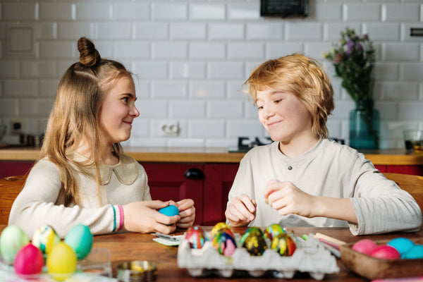 A young girl and boy painting easter eggs. Both are blond and facing each other while smiling. There are several painted eggs on the wooden table. A white brick wall is behind them.