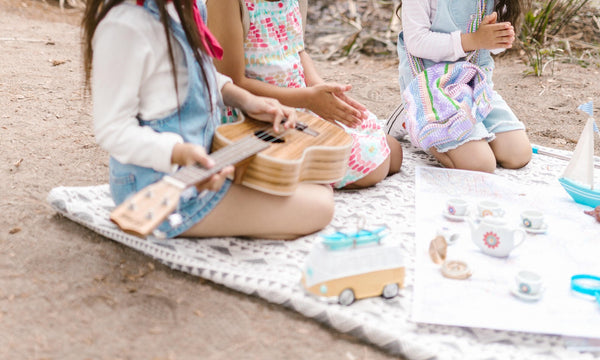 Three young girls (can't see their face) are sitting on a blanket at the beach having a tea party. One girl is holding a guitar. All three are wearing pastel colours. There is a toy truck on the blanket and a miniature tea party set.